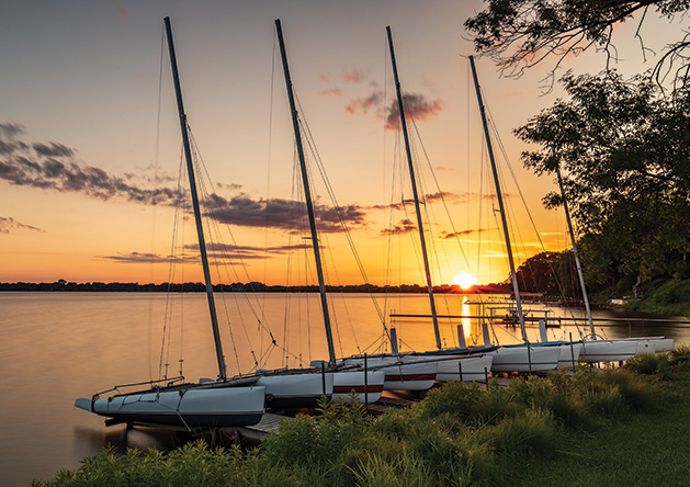 A row of catamarans float on White Bear Lake as the sun sets in the background.