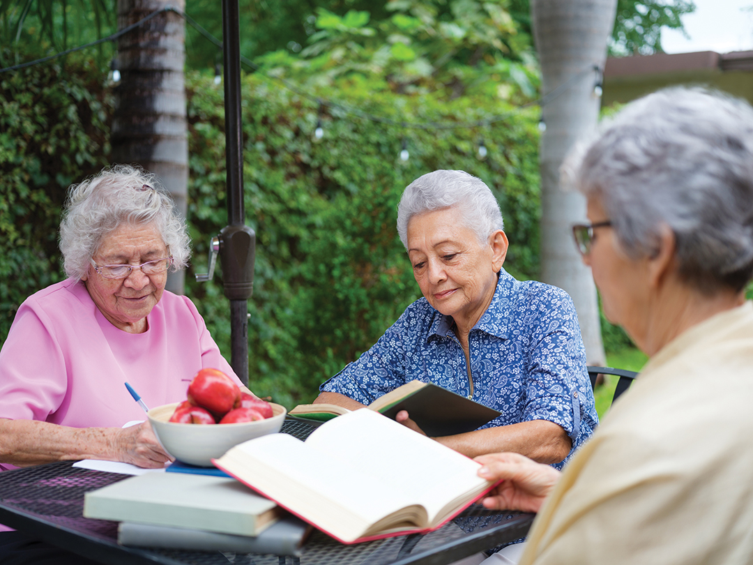 Three latin elderly women sitting on the patio at a table and reading books together.