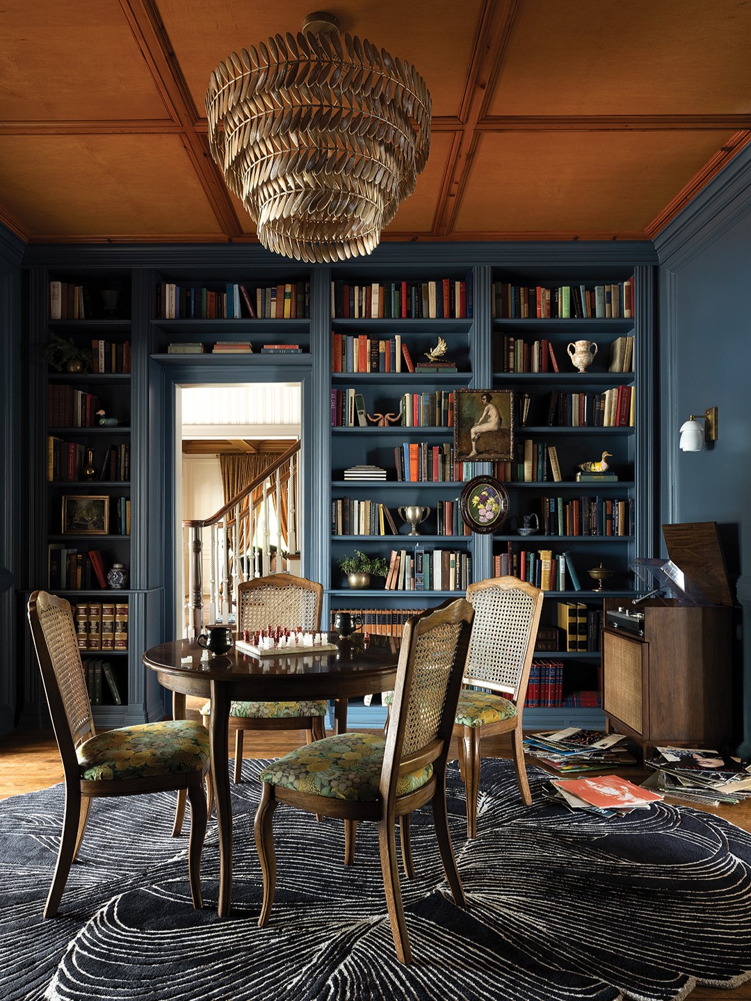 Blue Pencil Collective worked to preserve and foster a distinctive experience in each room. Custom details, carried through each space, bring comfort and warmth to the 6,650-square-foot home. Scalloped and rounded edges feature prominently from pendant lights and ceiling trim to rugs and furnishings. Patterns and textures are repeated in the wallpaper and curtains. In the library, pictured here, the scalloped rug, sourced in Italy, blends beautifully with an Art Deco lighting fixture and deep blue built-ins.